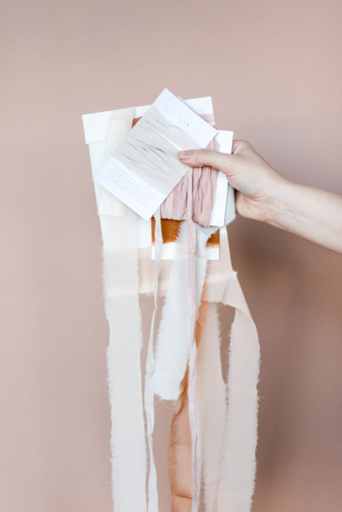 A hand holds up an array of neutral colored ribbons in front of a pastel background.