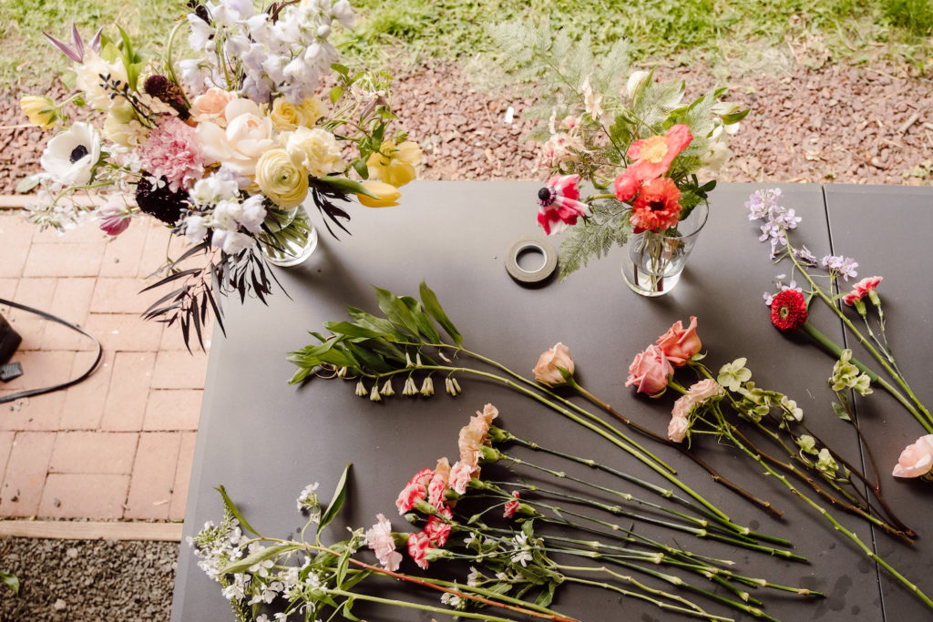 Flat-lay of various mixed flowers laid on the table, along with a two bunches of flower arrangements in clear vases photographed at Bouquet Bootcamp by @karenobrist