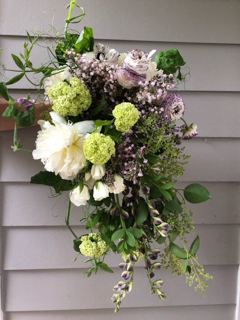 Elegant floral arrangement with white, purple, and green flowers, featuring various textures and greenery, held against a light grey background. Designed by The Floral Coach®, Amy Balsters.